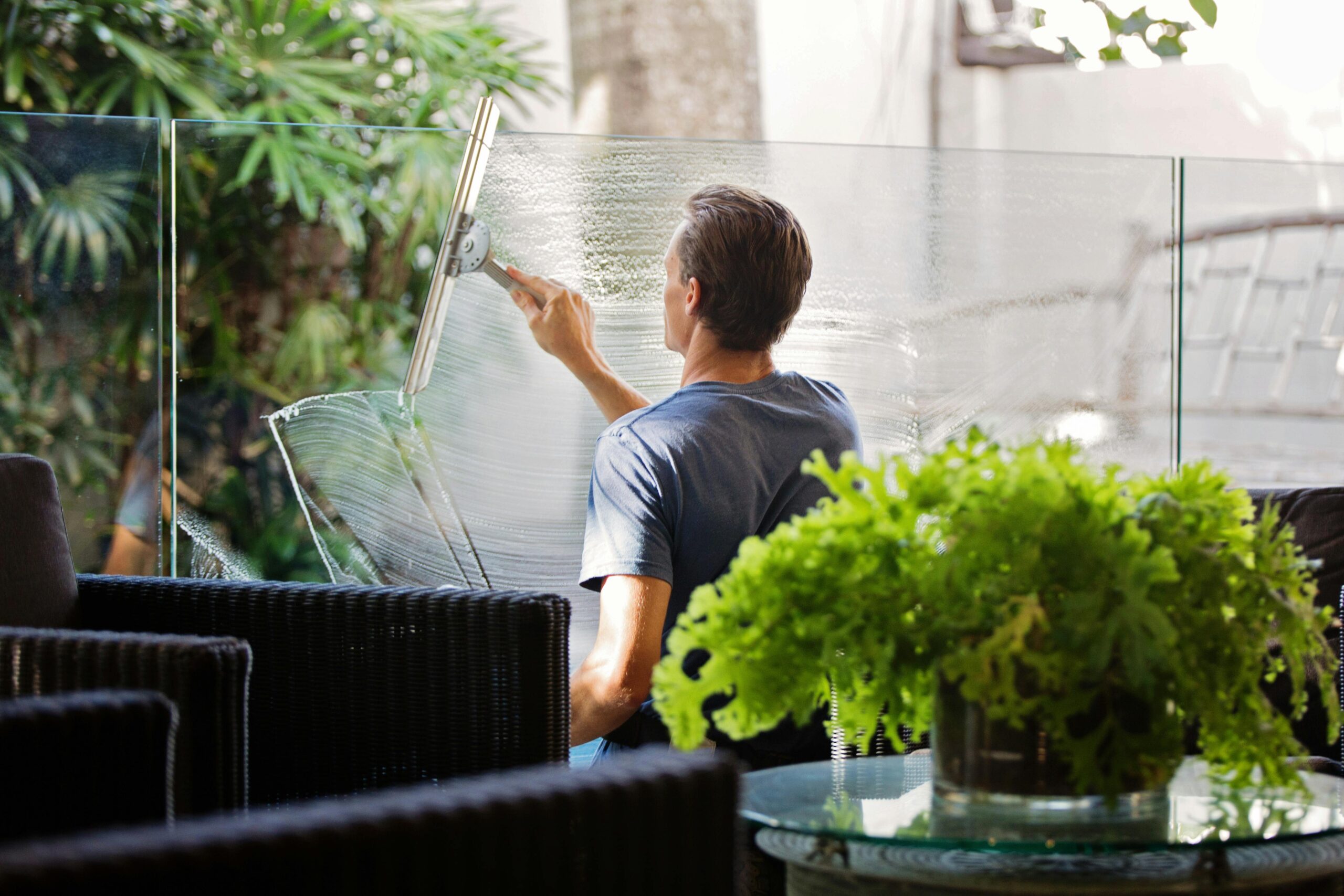 Cleaning services with a man cleaning a glass barrier outdoors with a squeegee, surrounded by greenery.