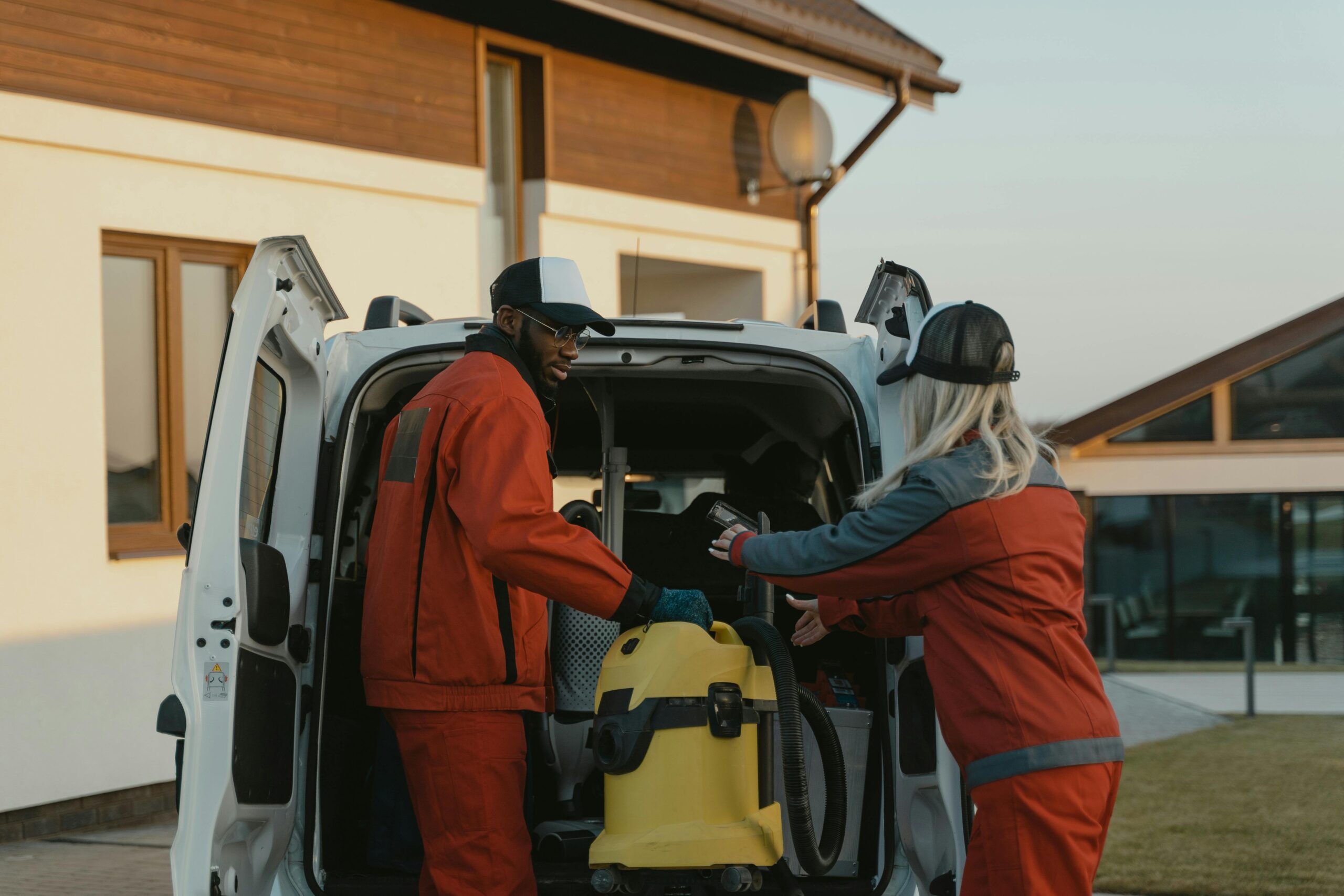 Cleaning services team in red uniforms load a vacuum into a white van outdoors.